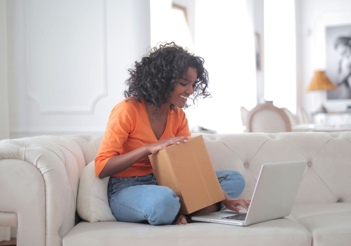 Une jeune femme souriante, assise sur un canapé, ouvre un colis en carton tout en consultant son ordinateur portable