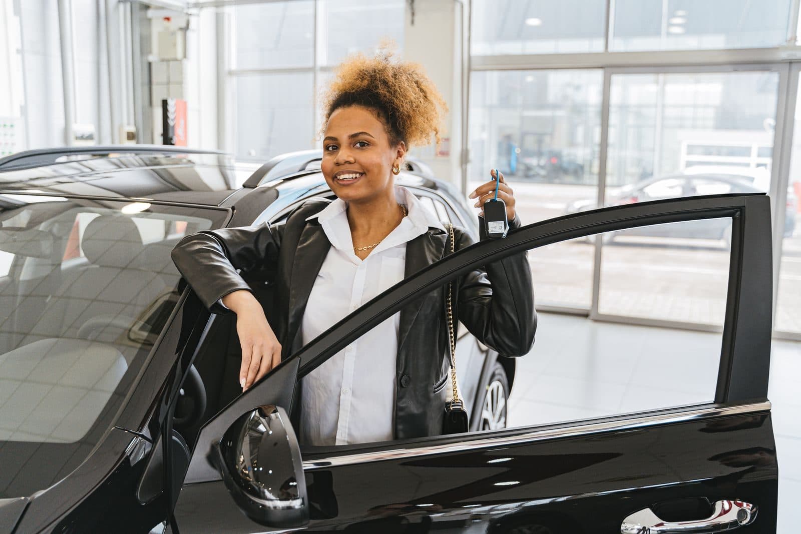 Une jeune femme souriante, une clé de voiture à la main, s’appuie sur la portière ouverte d’une berline noire dans une concession lumineuse