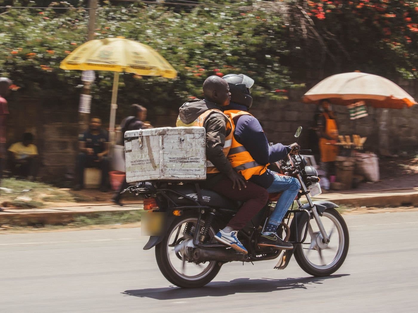 Deux hommes en gilet orange sur une moto, une grande caisse blanche fixée à l’arrière, roulent dans une rue de Nairobi, au Kenya, devant des étals ombragés de parasols